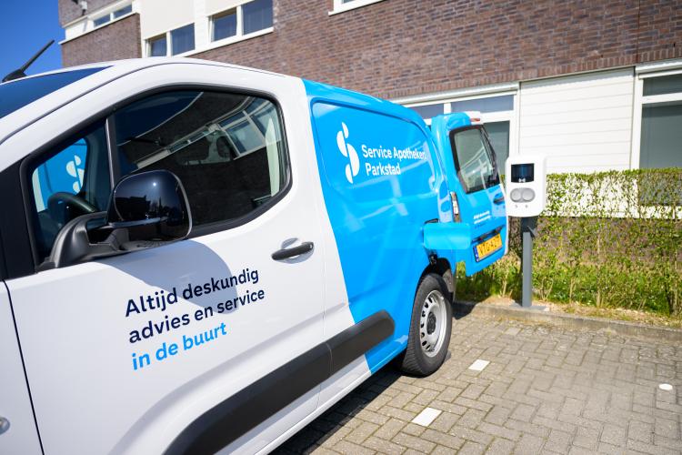 Witte en blauwe bestelwagen van Service Apotheek Parkstad geparkeerd voor een gebouw, met de tekst 'Altijd deskundig advies en service in de buurt' op de zijkant. Naast de wagen staat een laadpaal voor elektrische voertuigen.
