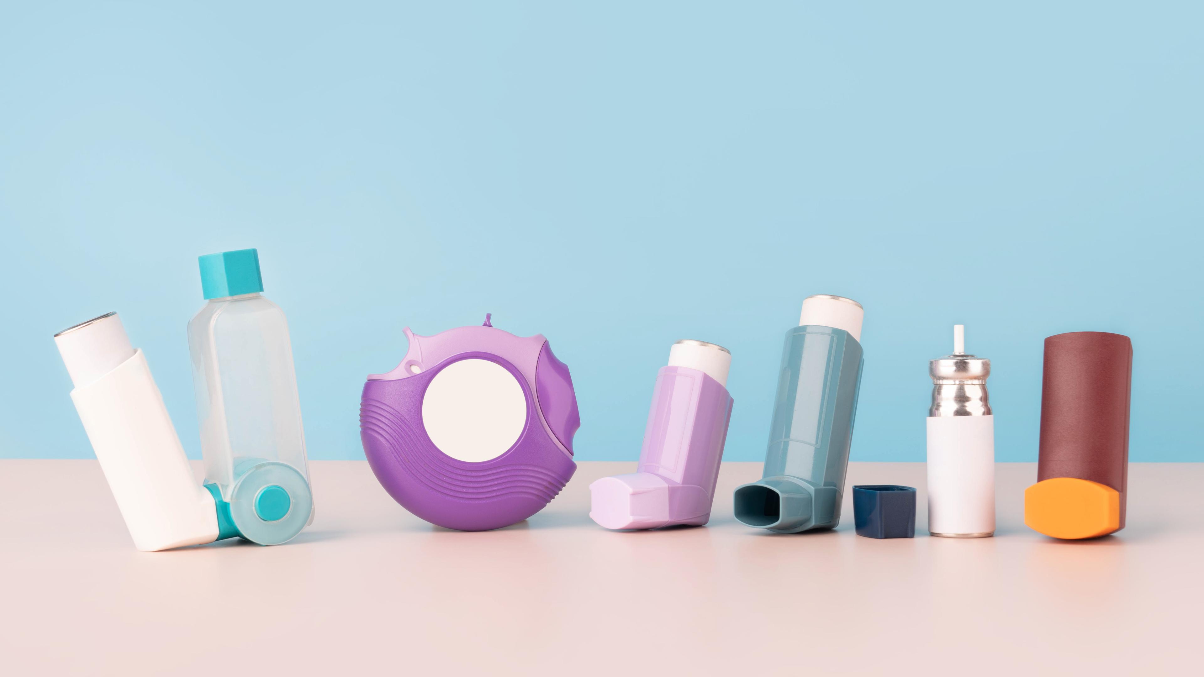 Een rij van zeven verschillende inhalers en medische hulpmiddelen op een lichtblauwe ondergrond. De apparaten variëren in kleur, waaronder wit, turquoise, paars, roze, blauw, zilver en bruin, en omvatten onder andere een astma-spacer.